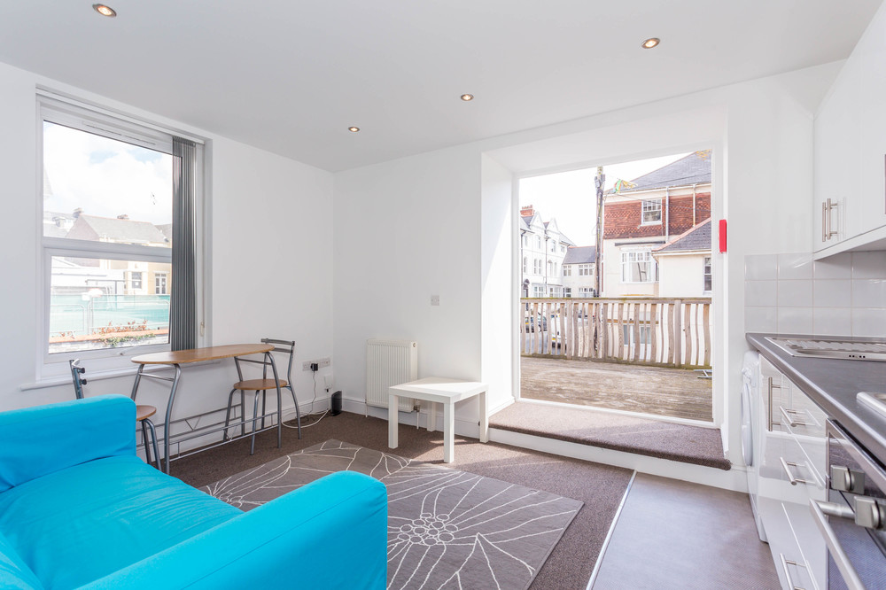 Living area and kitchen with blue two-seater sofa, dining table with two chairs and access to balcony