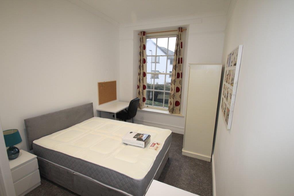 student bedroom with double bed, white desk with black chair, pinboard above desk on wall, two door wardrobe, bedside table with lamp