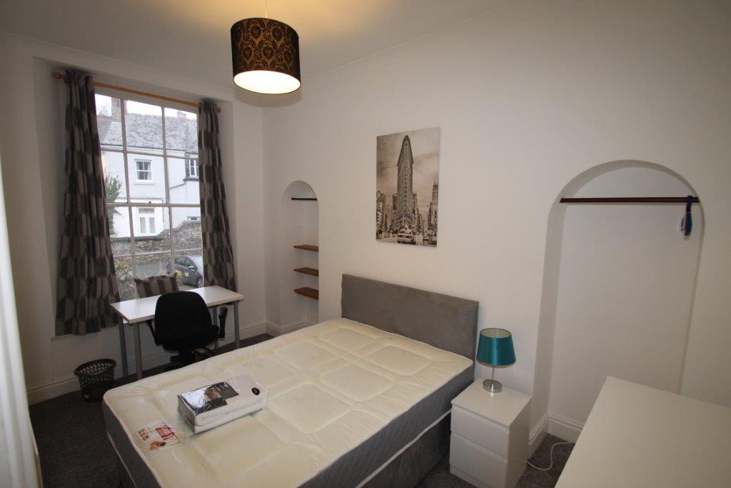 student bedroom with double bed, chest of drawers, white bedside table, desk with black office chair, shelves and clothing rail