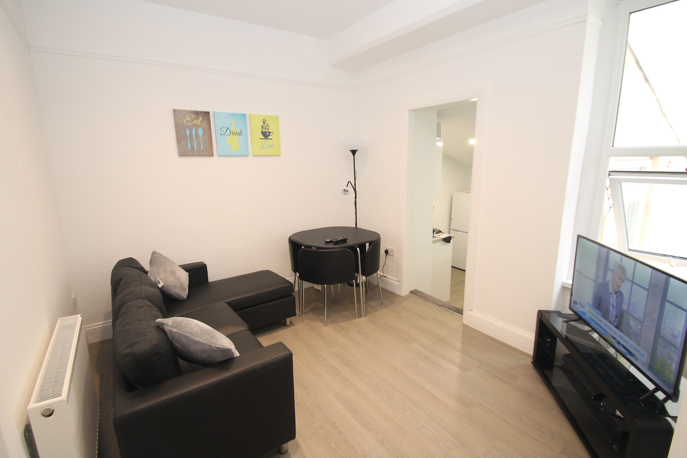 Living area with black L shaped sofa, small circular dining table, tv and stairwell leading to kitchen