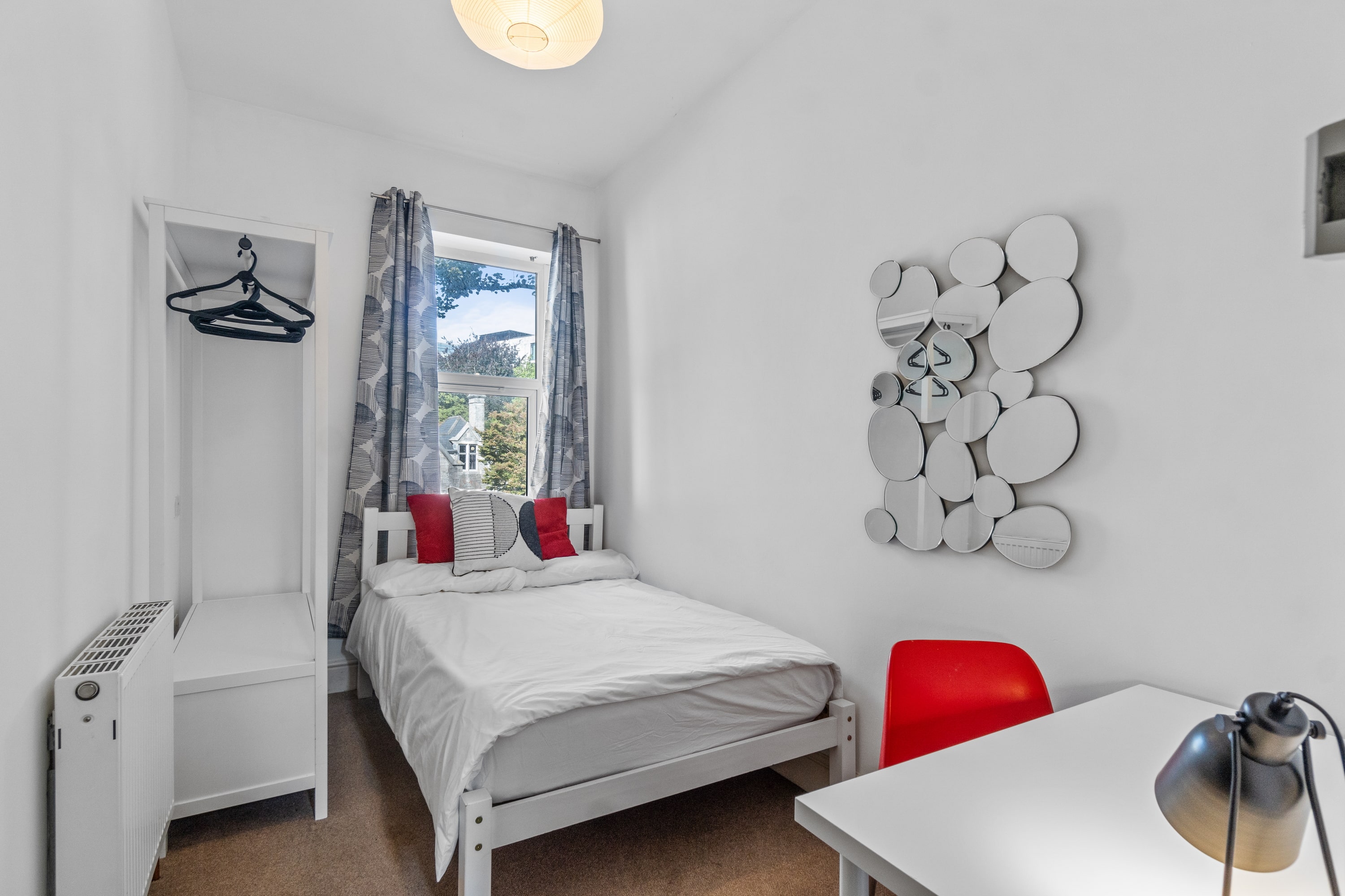 Student room with double bed, white desk with red chair, clothes rail with drawers and mirror