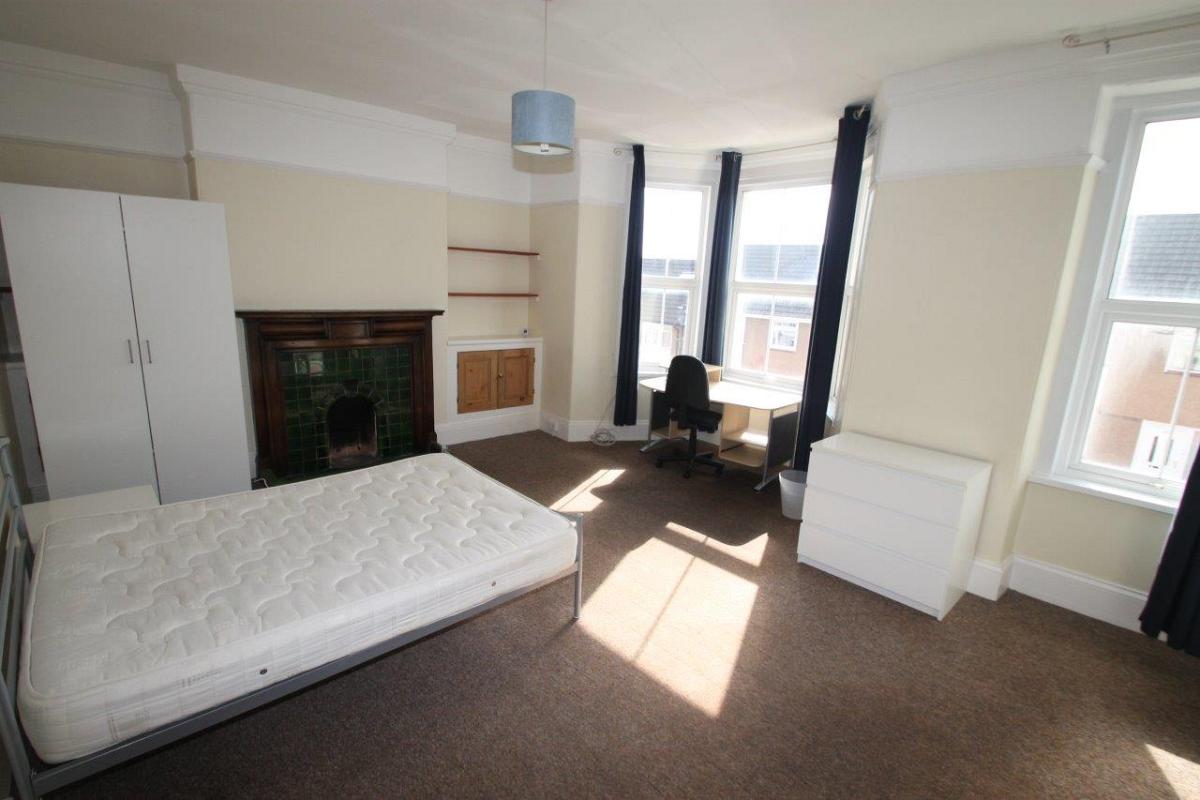student bedroom with double bed, two door wardrobe, chest of drawers, shelving unit and desk with office chair in front of large bay windows