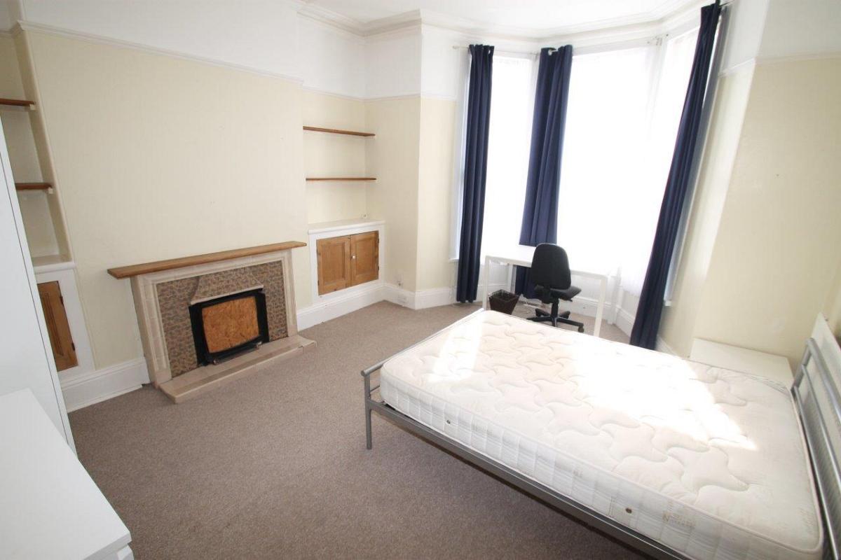 student bedroom with double bed, two door white wardrobe, four shelves and desk with black office chair in front of large bay windows