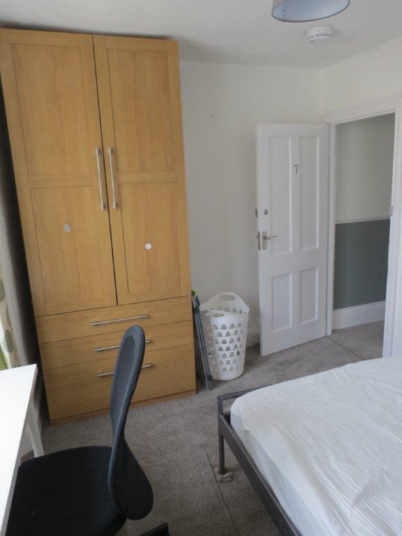 student bedroom with double bed, two door wardrobe with three drawers underneath, and desk with black office chair in front of window