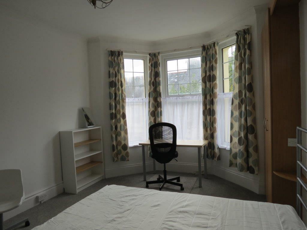 student bedroom with double bed, desk with black office chair in front of large bay windows, shelving unit and two door wardrobe