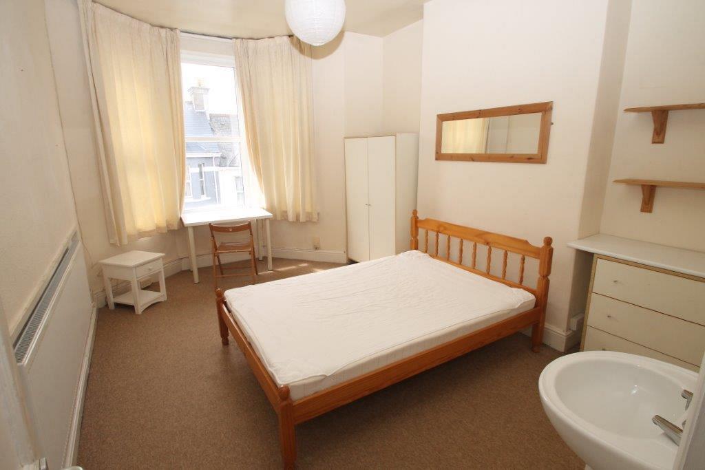 First student bedroom with double bed, two door white wardrobe, chest of drawers, bedside table, desk with chair, shelving above chest of drawers and sink