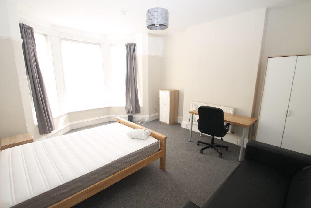 First student bedroom with double bed, two door wardrobe, tall chest of drawers, desk with black office chair, bedside table and black two-seater sofa