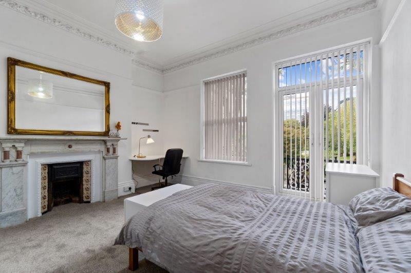 student bedroom with double bed, desk with black office chair, white chest of drawers and large mirror above feature fireplace