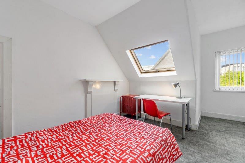 student bedroom with double bed, bedside table, white desk with red chair in front of skylight  