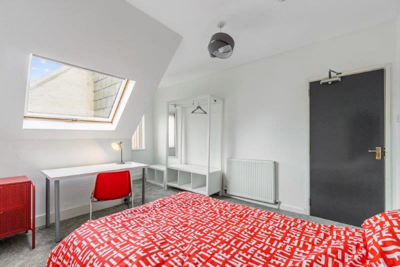 student bedroom with double bed, bedside table, white desk with red chair and white clothing rail with storage underneath