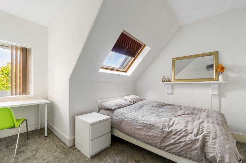 student bedroom with doubled bed, bedside table, desk in front of window with green chair and mirror above fireplace