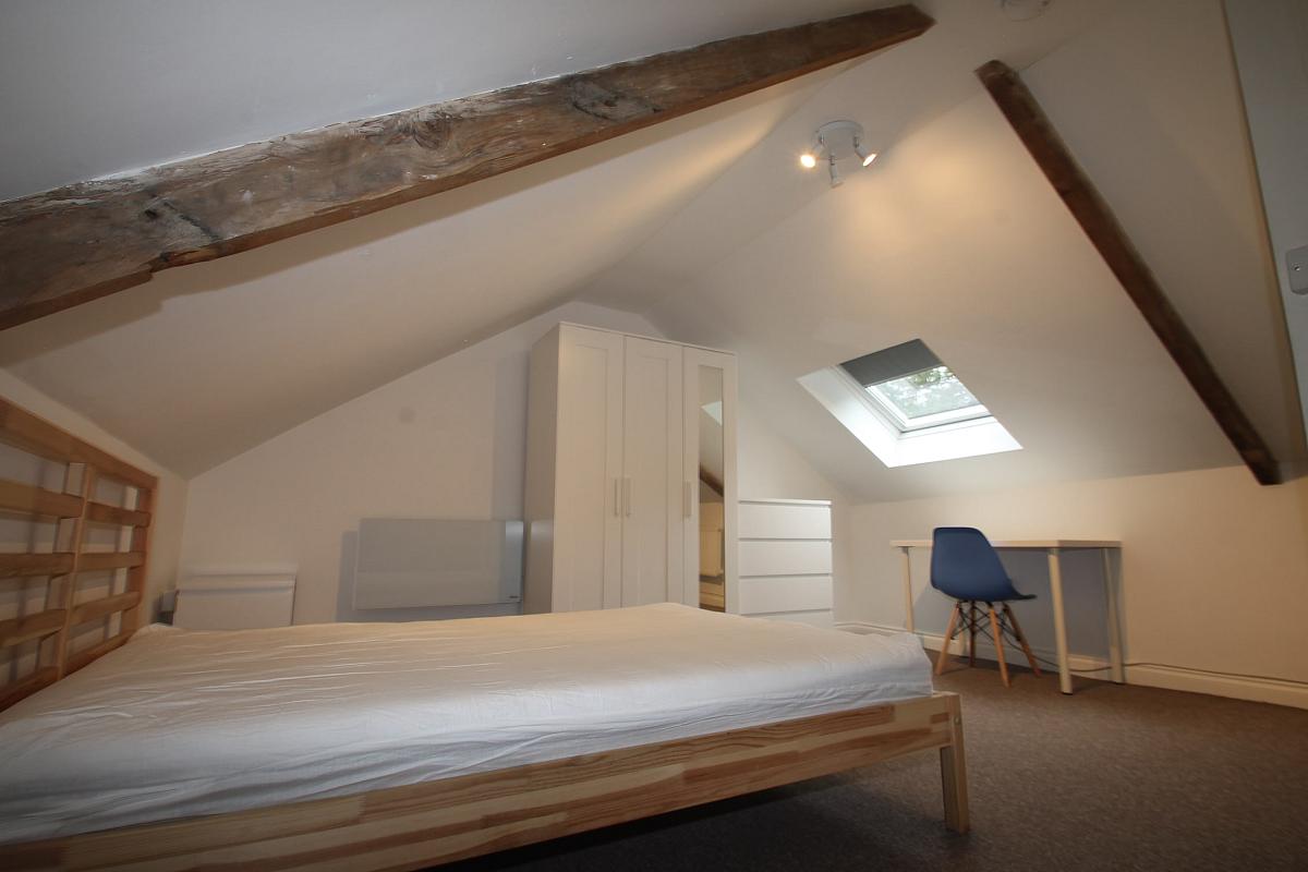 Sixth student bedroom with double bed, three door wardrobe, white chest of drawers, desk with blue chair, room has exposed wooden beams and skylight