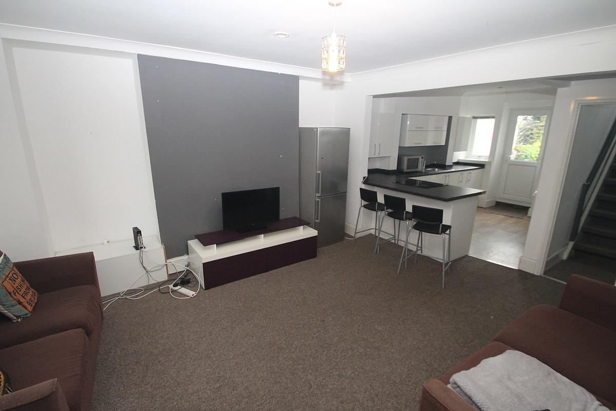 spacious open plan kitchen and living area with fridge freezer, breakfast bar with three black bar stools, two brown two-seater sofas, large bay windows, grey feature wall and tv on tv stand