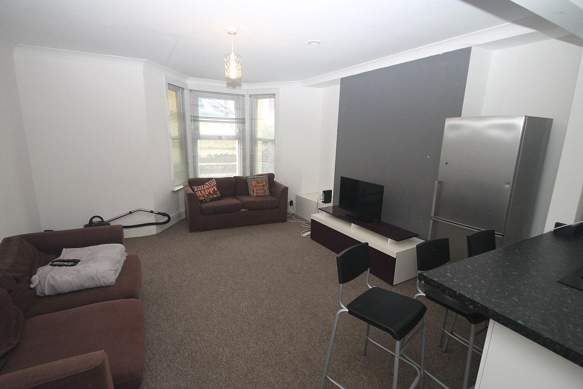 open plan kitchen and living area with fridge freezer, breakfast bar with three black bar stools, two brown two-seater sofas, large bay windows, grey feature wall and tv on tv stand