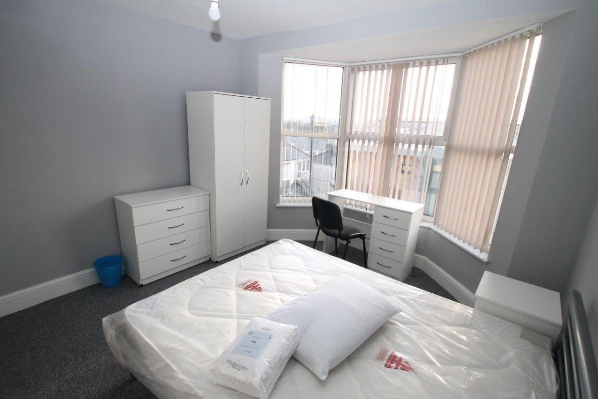 Grey walled student bedroom with double bed, two door wardrobe, chest of drawers and desk in front of large bay window