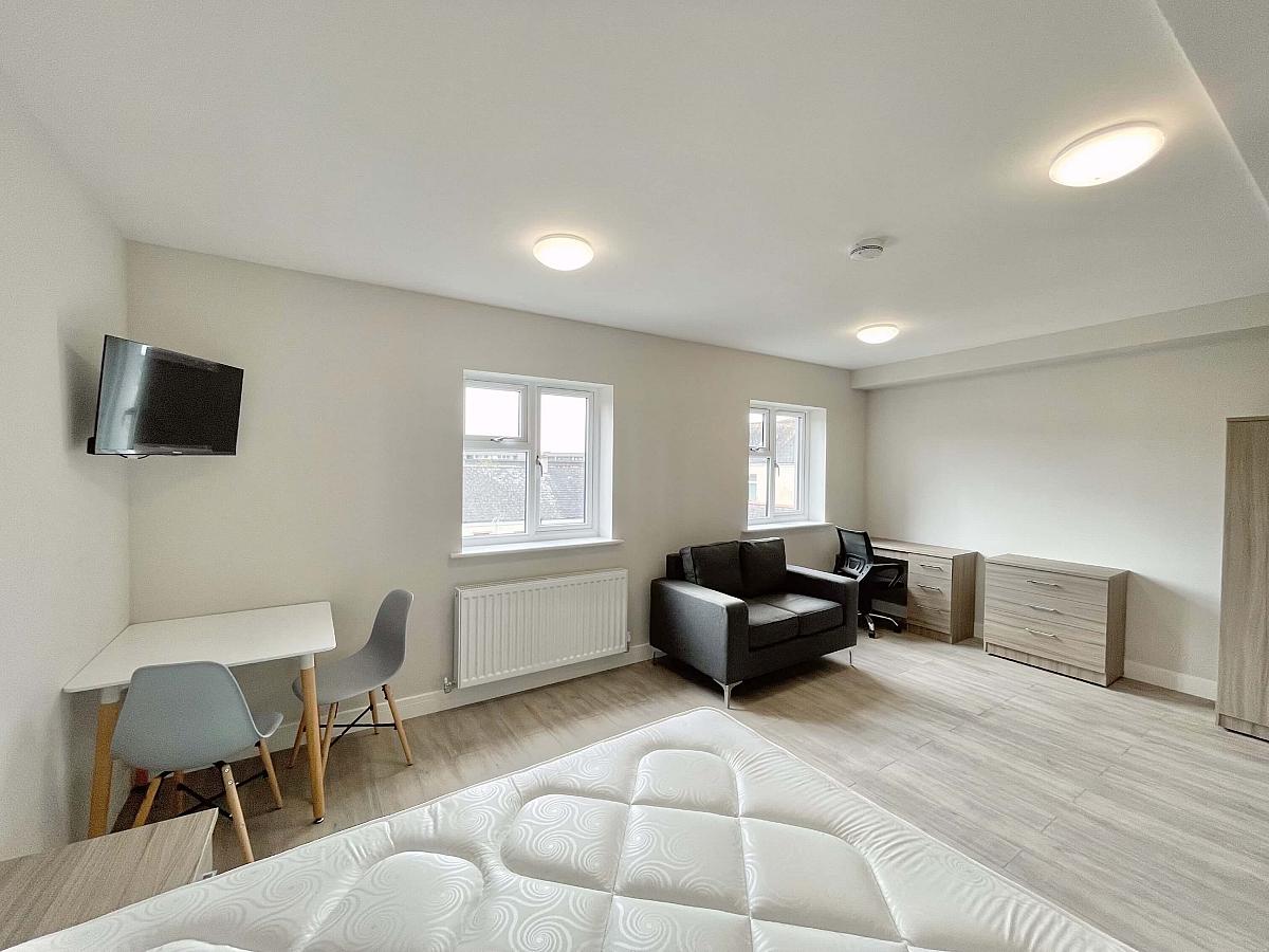 Living space with desk, chest of drawers, two door wardrobe, large grey arm chair, dining table with two chairs and wall mounted tv