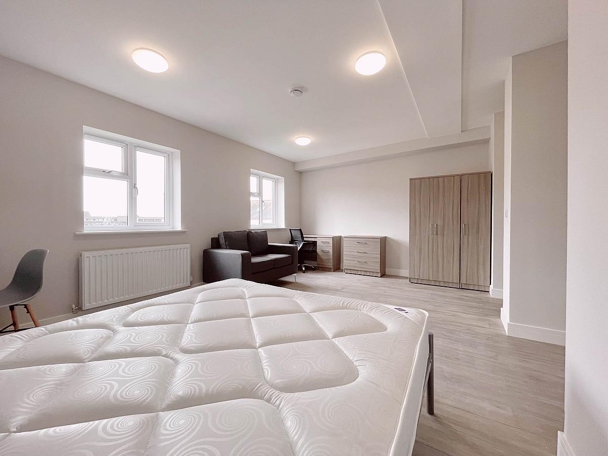 Living space with double bed, three door wardrobe, desk, chest of drawers, large grey arm chair with two large windows