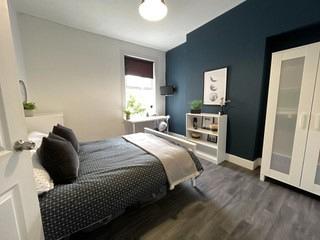 student bedroom with double bed, bedside table, two door wardrobe, white desk with chair and white shelving unit
