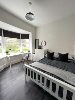 student bedroom with double bed, desk with black chair in front of large bay windows, white bedside table and chest of drawers