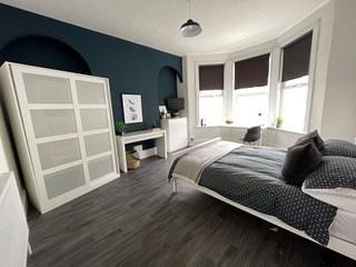 student bedroom with double bed, white two door wardrobe, desk, white chest of drawers and large bay windows