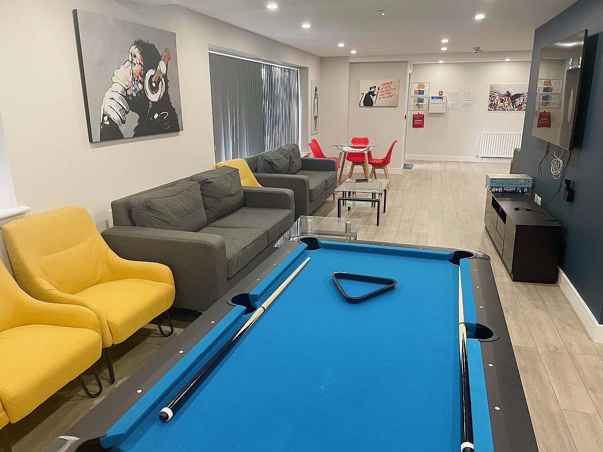 Downstairs communal area with two double sofas, three yellow arm chairs, wall mounted tv and pool table