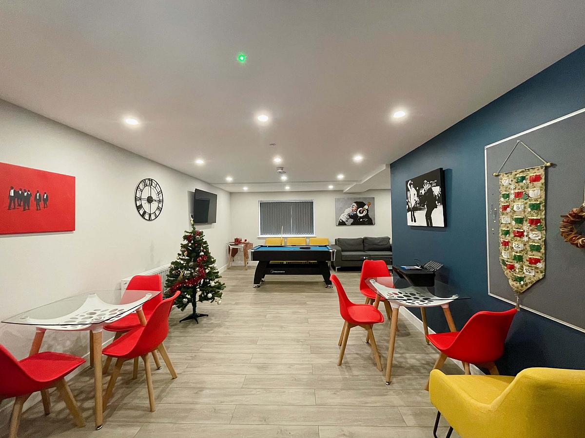 Downstairs communal area with two tables with seating, pool table, wall mounted tv, two double sofas and three yellow armchairs 