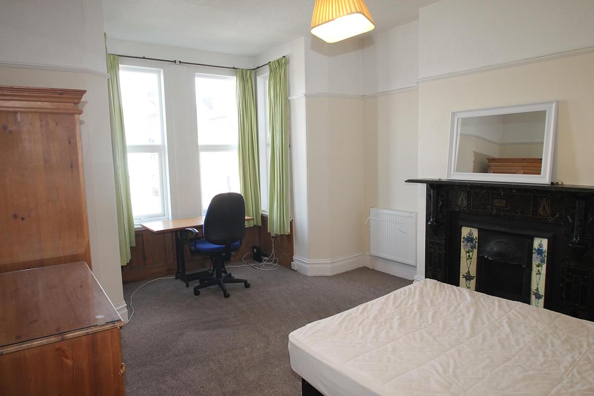 first student bedroom with double bed, two door wardrobe, desk with blue office chair in front of bay windows, chest of drawers and fireplace