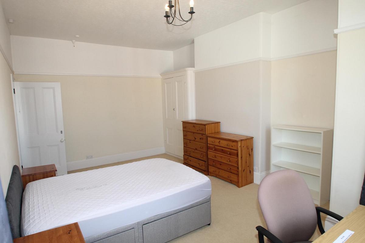 Fifth student bedroom with double bed, built-in wardrobe, two chest of drawers, shelving unit, bedside table and desk with chair in front of windows