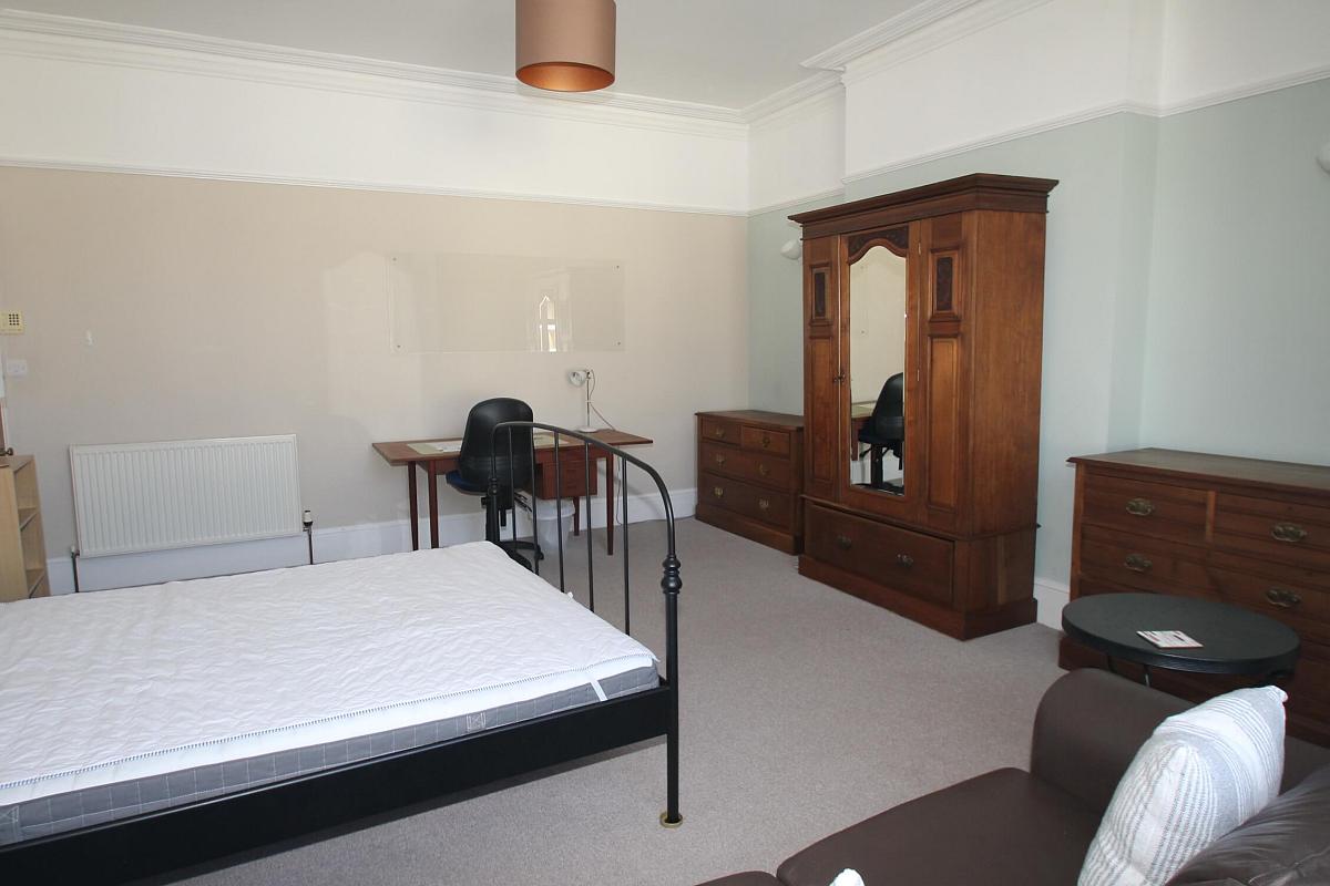 fifth student bedroom with double bed, two chests of drawers, bedside table, black leather two-seater sofa, desk with blue office chair