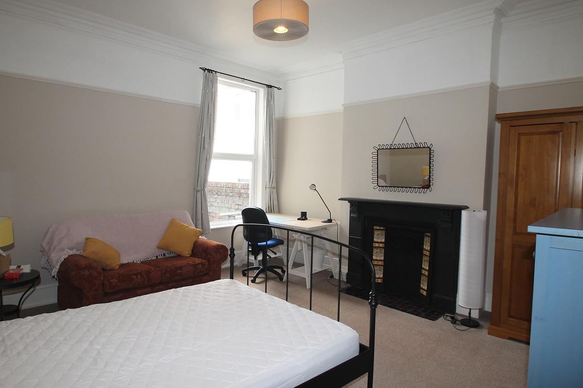first student room with double bed, bedside table, two-seater sofa, blue chest of drawers, two door wardrobe, desk with blue office chair and mirror mounted above black fireplace