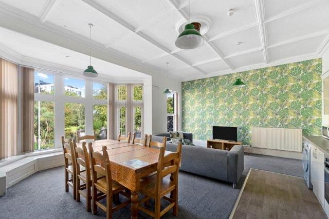 Spacious living and kitchen areas with extremely large bay windows, dining table with eight dining chairs, two grey sofas, tv on tv stand