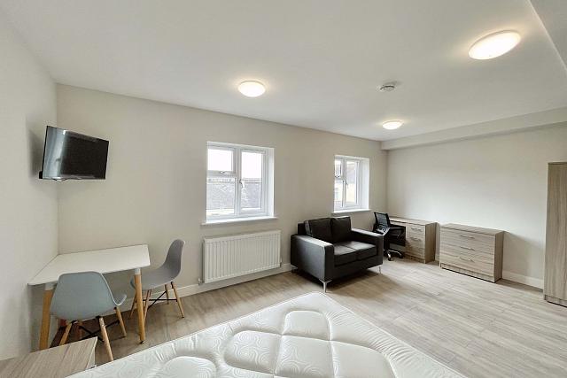 Living space with desk, chest of drawers, two door wardrobe, large grey arm chair, dining table with two chairs and wall mounted tv