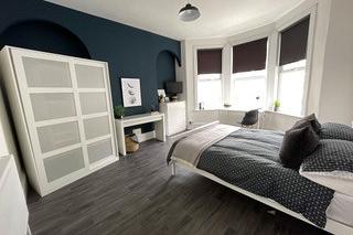 student bedroom with double bed, white two door wardrobe, desk, white chest of drawers and large bay windows